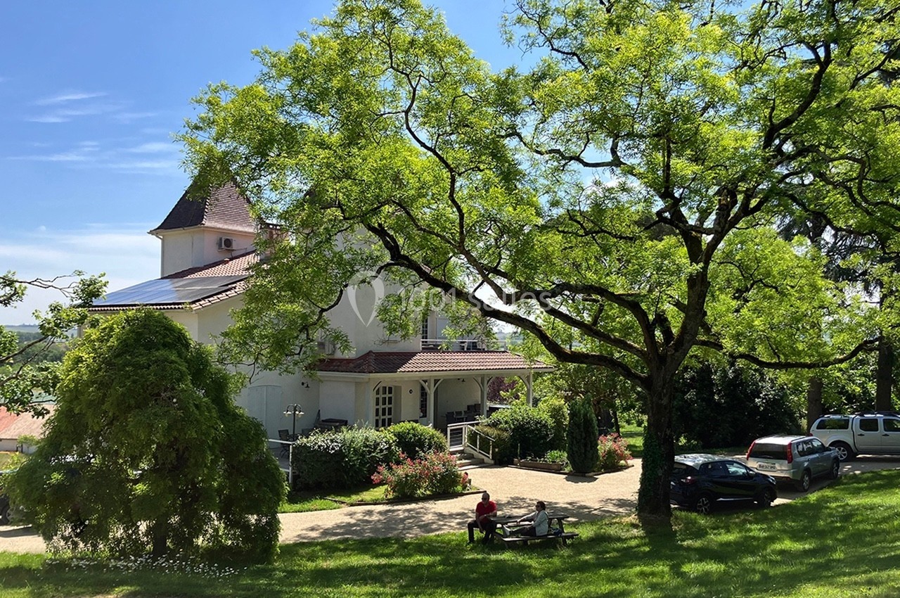 Bâtiment blanc entouré d'arbres et de verdure, avec des voitures garées et une personne assise sur un banc.