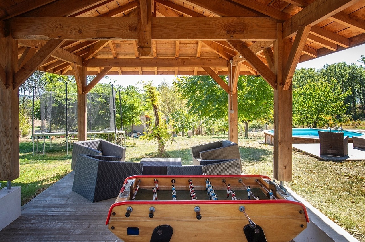 Terrasse couverte en bois avec baby-foot, salon de jardin, vue sur trampoline, piscine et espace vert.