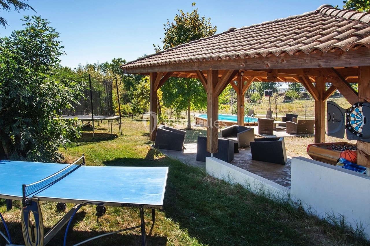 Espace extérieur avec terrasse couverte, salon de jardin, table de ping-pong, trampoline et piscine en arrière-plan.