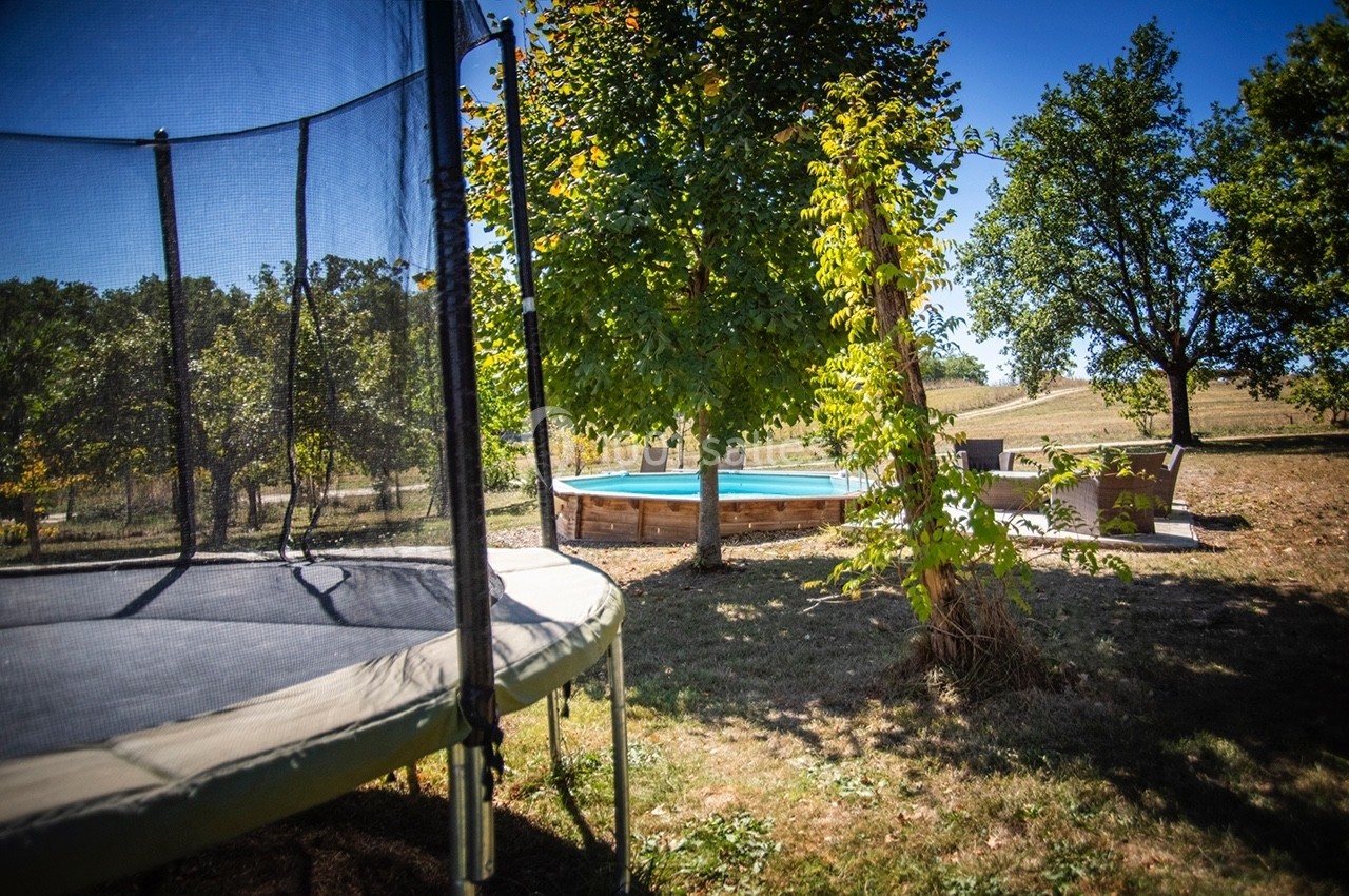 Trampoline et piscine extérieure entourés d'arbres dans un jardin ensoleillé.