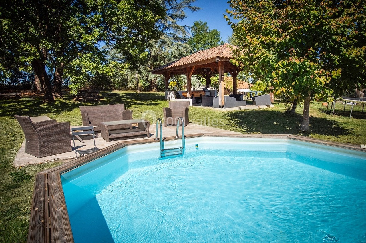 Piscine octogonale extérieure entourée de mobilier de jardin et d'arbres, avec un abri en bois au fond.