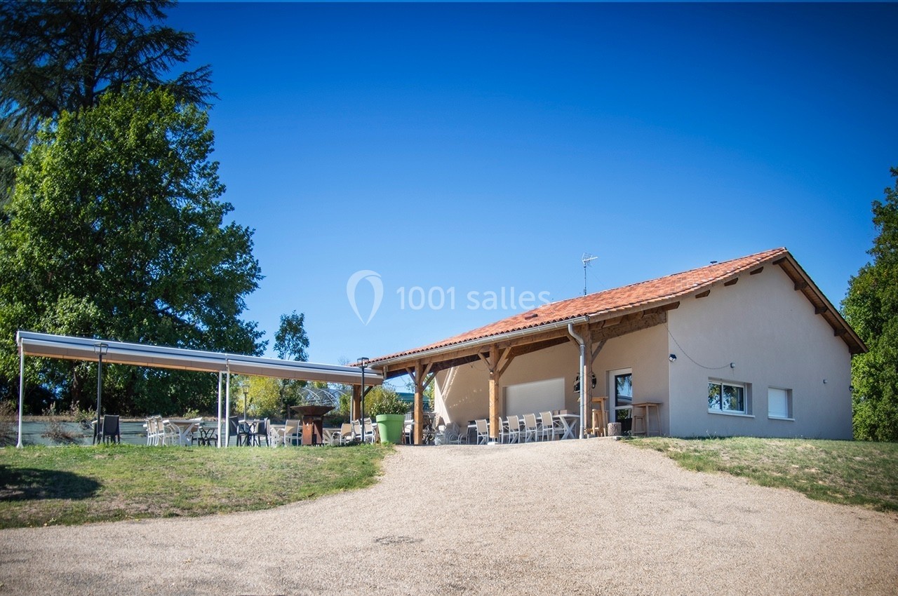 Bâtiment avec toit en tuiles, terrasse couverte et mobilier extérieur, entouré d'arbres sous un ciel bleu.