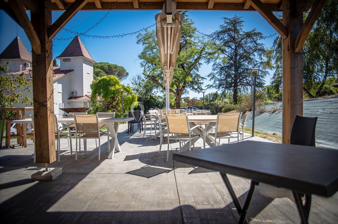 Terrasse extérieure ombragée avec tables et chaises, entourée de végétation et vue sur des bâtiments en arrière-plan.