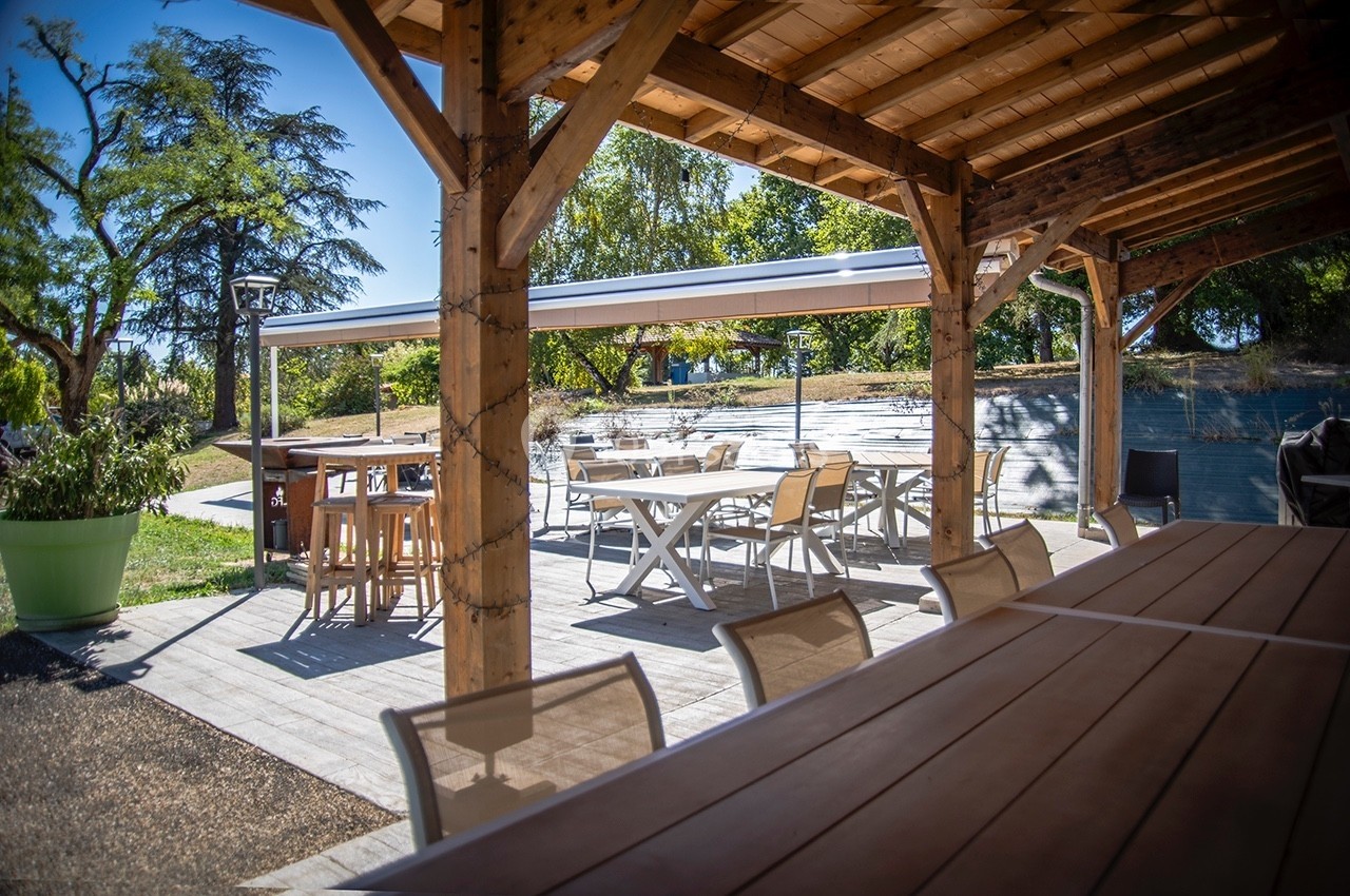 Terrasse extérieure ombragée avec tables et chaises en bois et métal, entourée de verdure et éclairée par la lumière…