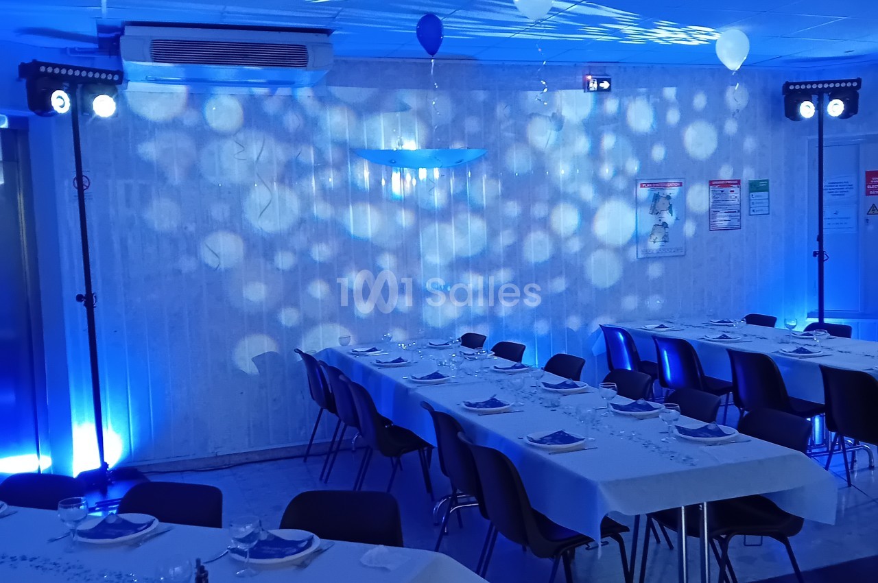 Salle intérieure dressée avec tables longues blanches, chaises noires, éclairage bleu ambiant et projecteurs - capacité environ 30-40 places.