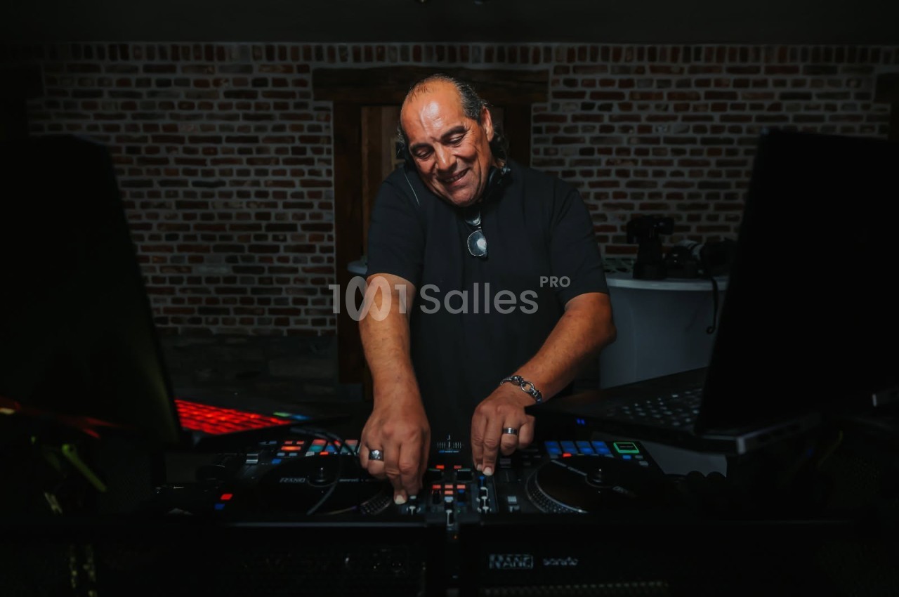 Un DJ souriant, vêtu d'un t-shirt noir, règle sa table de mixage en pleine lumière tamisée, devant un mur en briques.