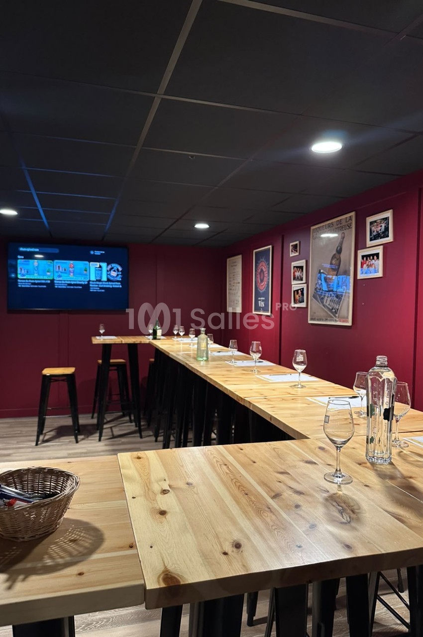 Salle de dégustation aux murs bordeaux avec de longues tables hautes en bois clair, verres à pied, un écran au mur et des tabourets hauts.