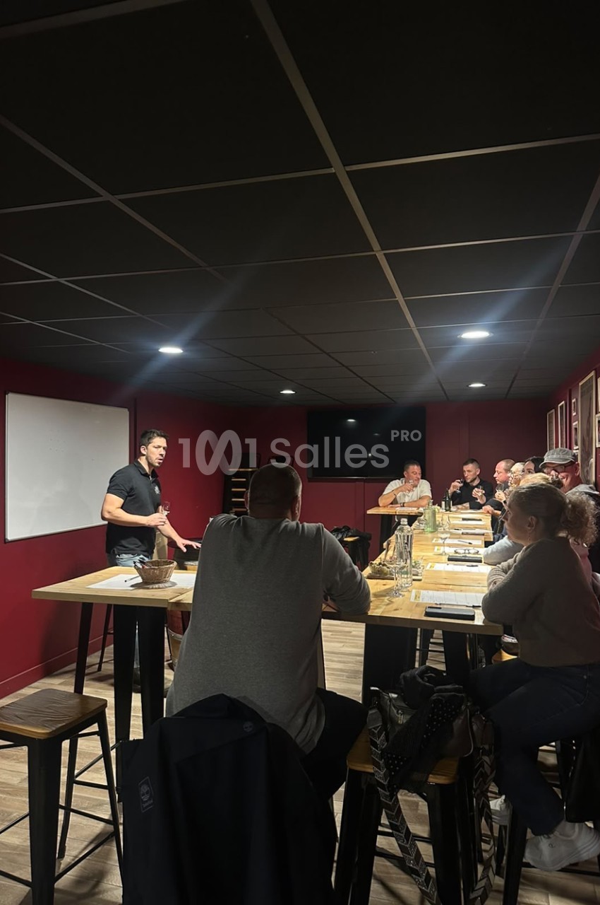 Salle aux murs bordeaux, un homme debout présente à des personnes assises autour de grandes tables hautes en bois.