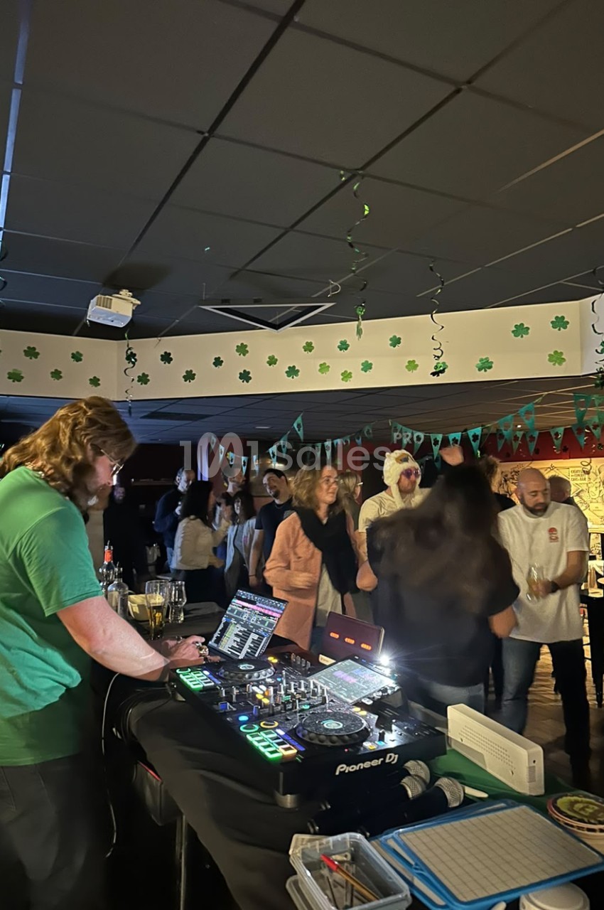 Un DJ en t-shirt vert derrière sa console de mixage, face à des convives debout dans une salle décorée de trèfles verts.