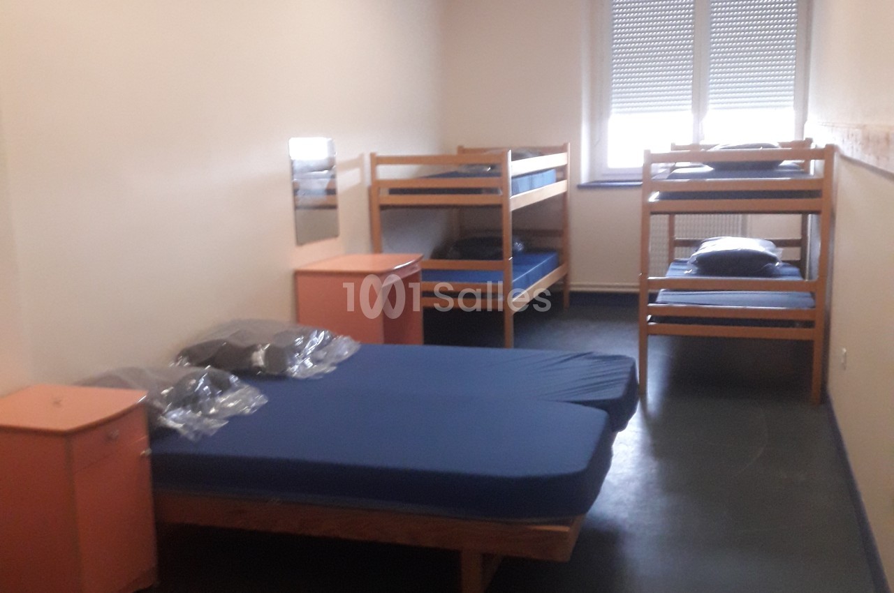 Chambre avec lit simple à matelas bleu au premier plan et deux lits superposés en bois contre les murs, fenêtres avec volets fermés.