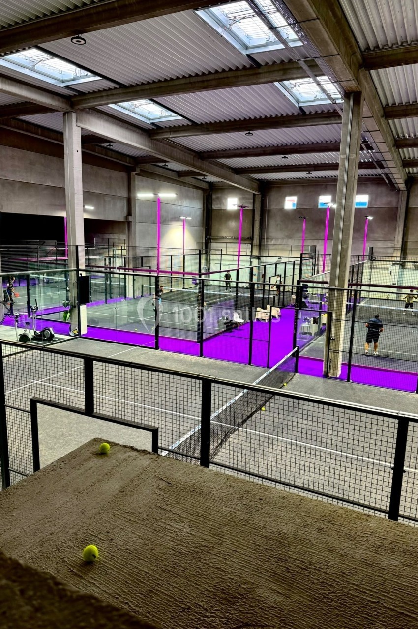 Vue intérieure d'un complexe sportif avec plusieurs terrains de padel entourés de grillages et éclairés par des néons…