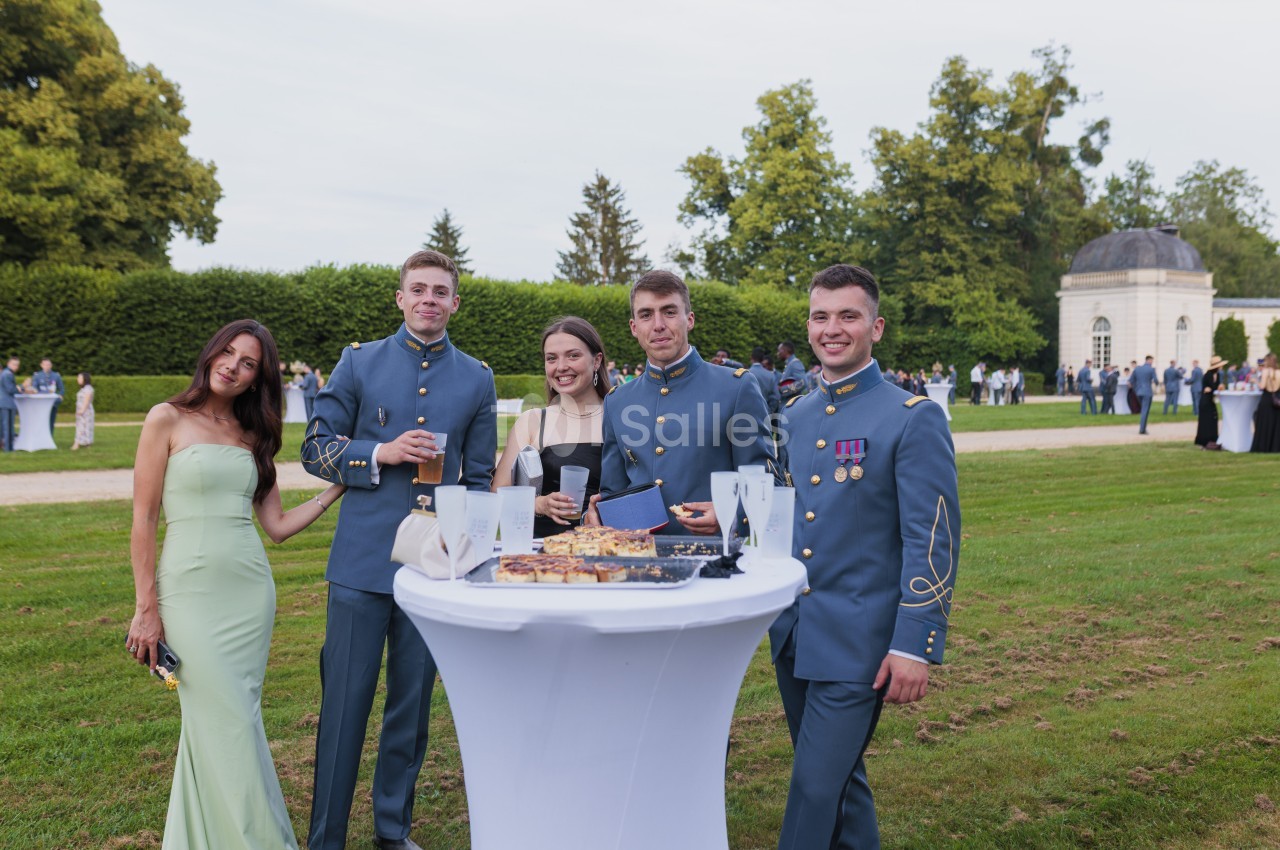 Cinq convives en tenue formelle autour d'un cocktail dressé en plein air, parc verdoyant et bâtiment classique en arrière-plan.