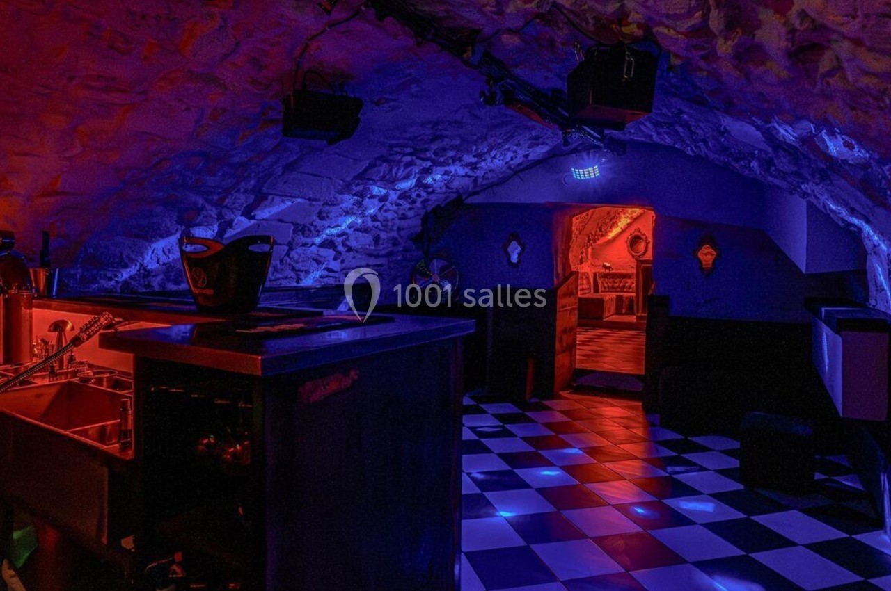 Salle voûtée avec éclairage tamisé rouge et bleu, sol en damier noir et blanc, bar et mobilier sombre.
