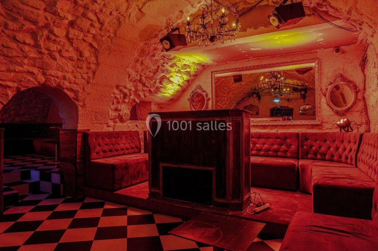 Salle voûtée en pierre avec banquettes rouges, mobilier en bois et sol à carreaux noirs et blancs.