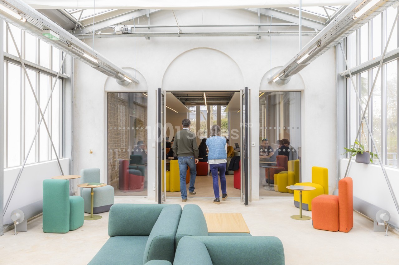 Espace lumineux aux murs blancs et verrière, avec canapés et fauteuils colorés (vert, jaune, orange) disposés au premier plan, et deux personnes debout devant une porte ouvrant sur une autre salle. Let me verify length: roughly 165 characters, too long.