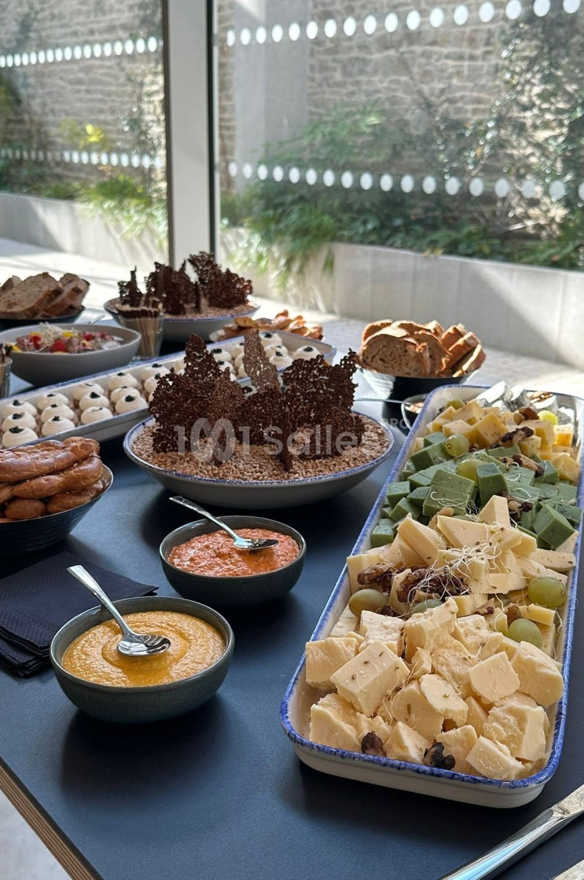 Buffet dressé sur table sombre : plateaux de fromages en cubes avec raisins et noix, bols de dips orangé et rouge, pains, bretzels et desserts en chocolat, près d'une baie vitrée.