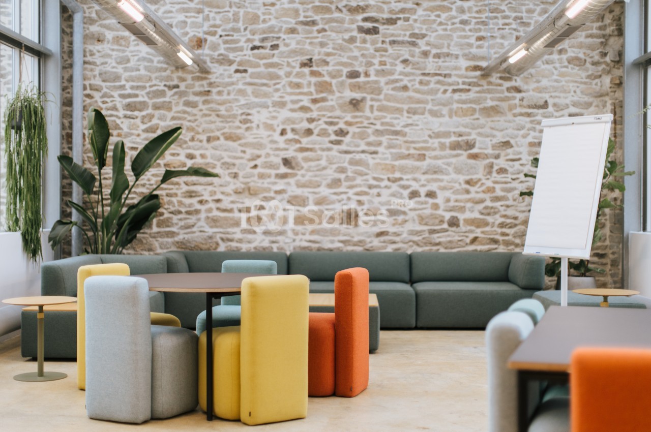 Espace lounge lumineux avec mur en pierre apparente, canapés gris-vert, poufs colorés jaunes et orange autour de tables basses, et paperboard vierge.
