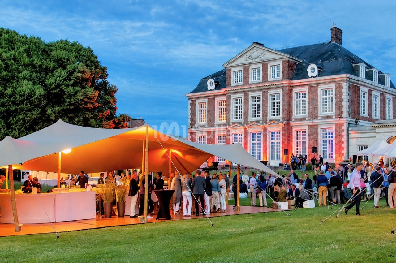 Tentes de réception orange et blanches illuminées en soirée devant château en briques, convives circulant sur pelouse.