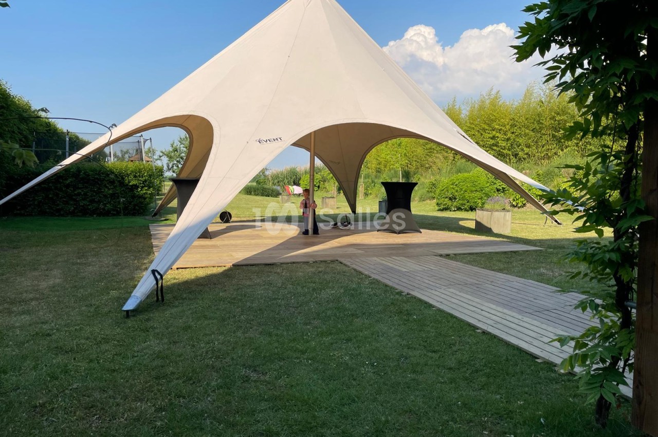 Tente stretch beige avec arches marron et terrasse bois dans jardin verdoyant.