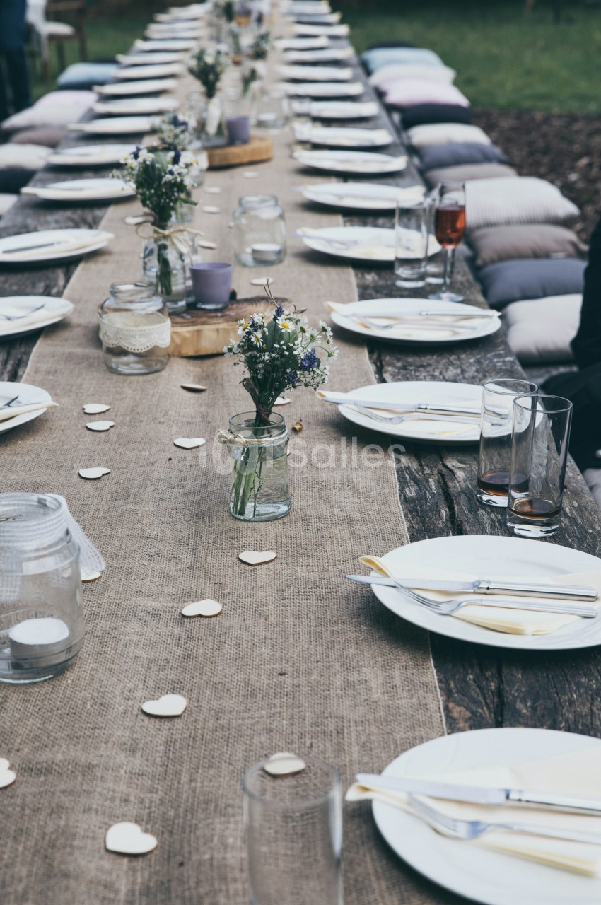 Table extérieure dressée en longueur avec assiettes blanches, verres et petits bouquets - coussins gris sur bancs.
