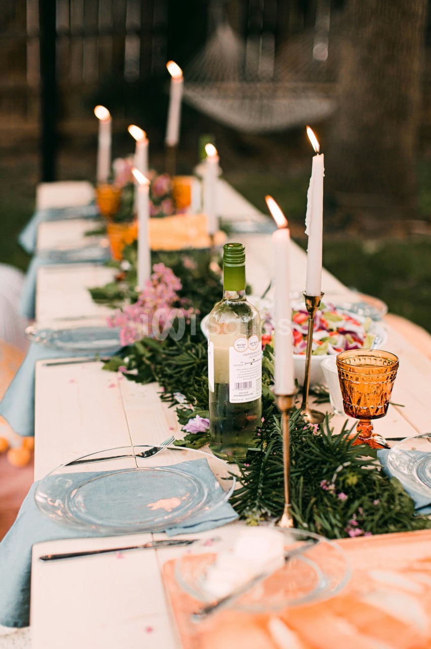 Table dressée en extérieur avec assiettes, couverts, chandeliers à bougies blanches, bouteille de vin et fleurs roses le long du chemin de table.
