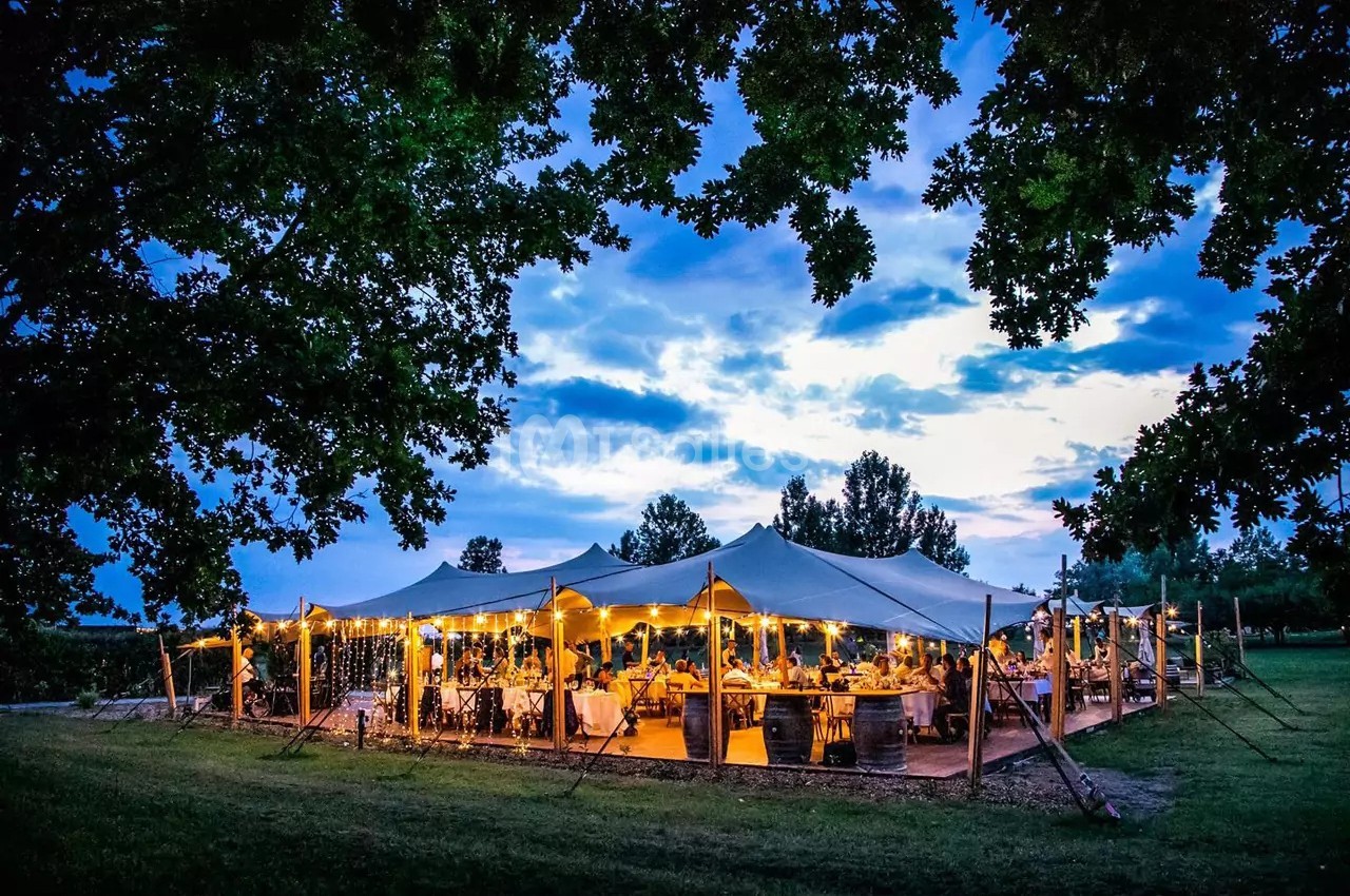 Chapiteau blanc illuminé en soirée avec tables dressées, convives attablés et ciel bleu nuageux en arrière-plan.