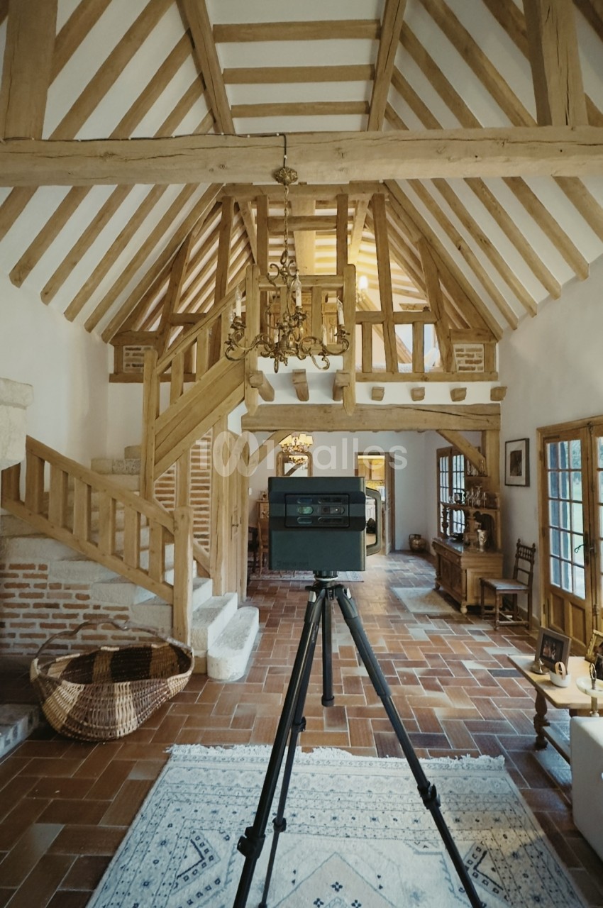 Intérieur en colombages avec charpente apparente, escalier en bois et caméra sur trépied.