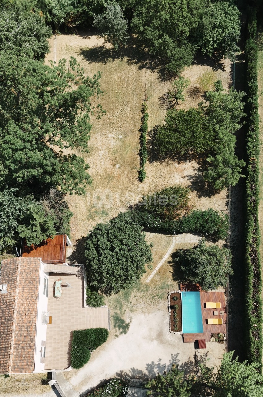 Propriété vue d'en haut avec piscine turquoise, terrasse en bois et bâtisse blanche entourée d'arbres.