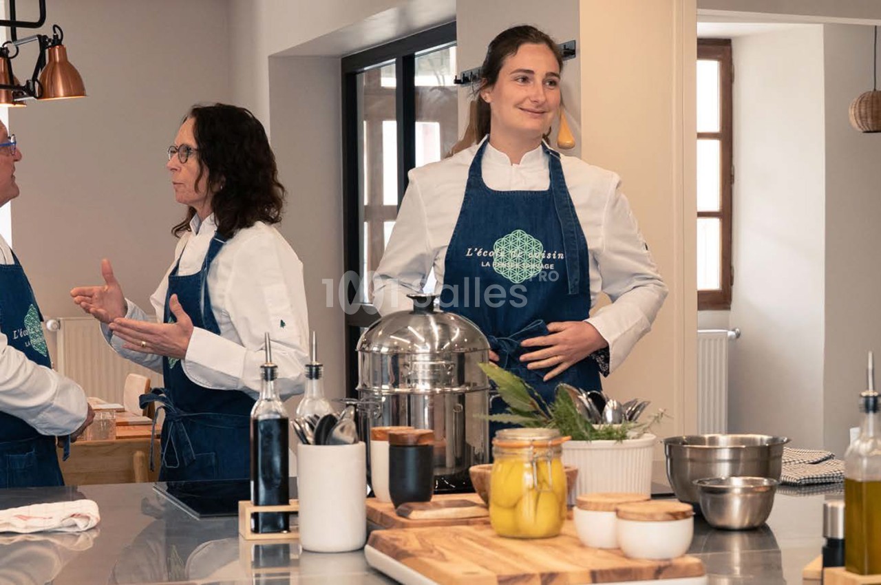 Deux femmes en tablier bleu devant un plan de travail équipé d'ustensiles de cuisine et d'ingrédients en bocal.