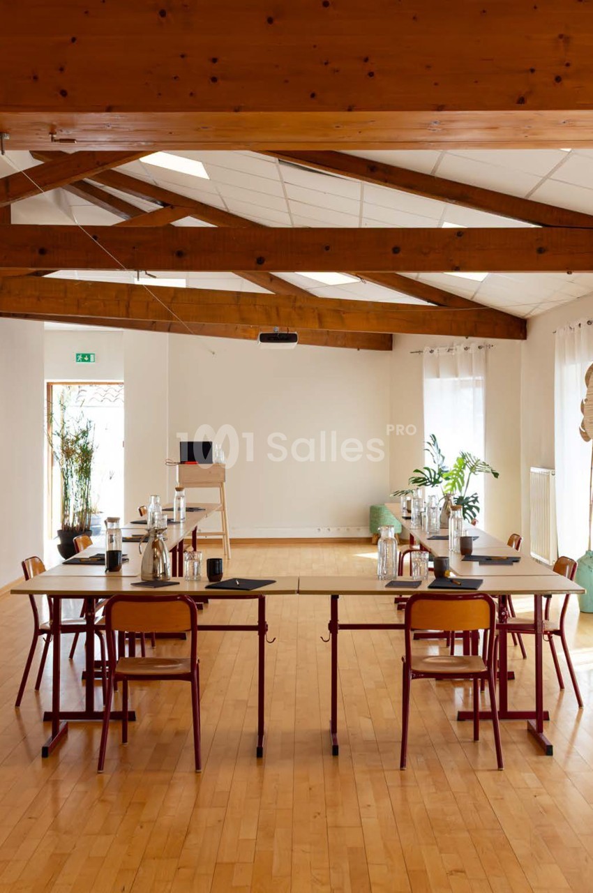 Salle de réunion avec poutres en bois, chaises rouges autour d'une table centrale, parquet clair, baies vitrées et plantes vertes.