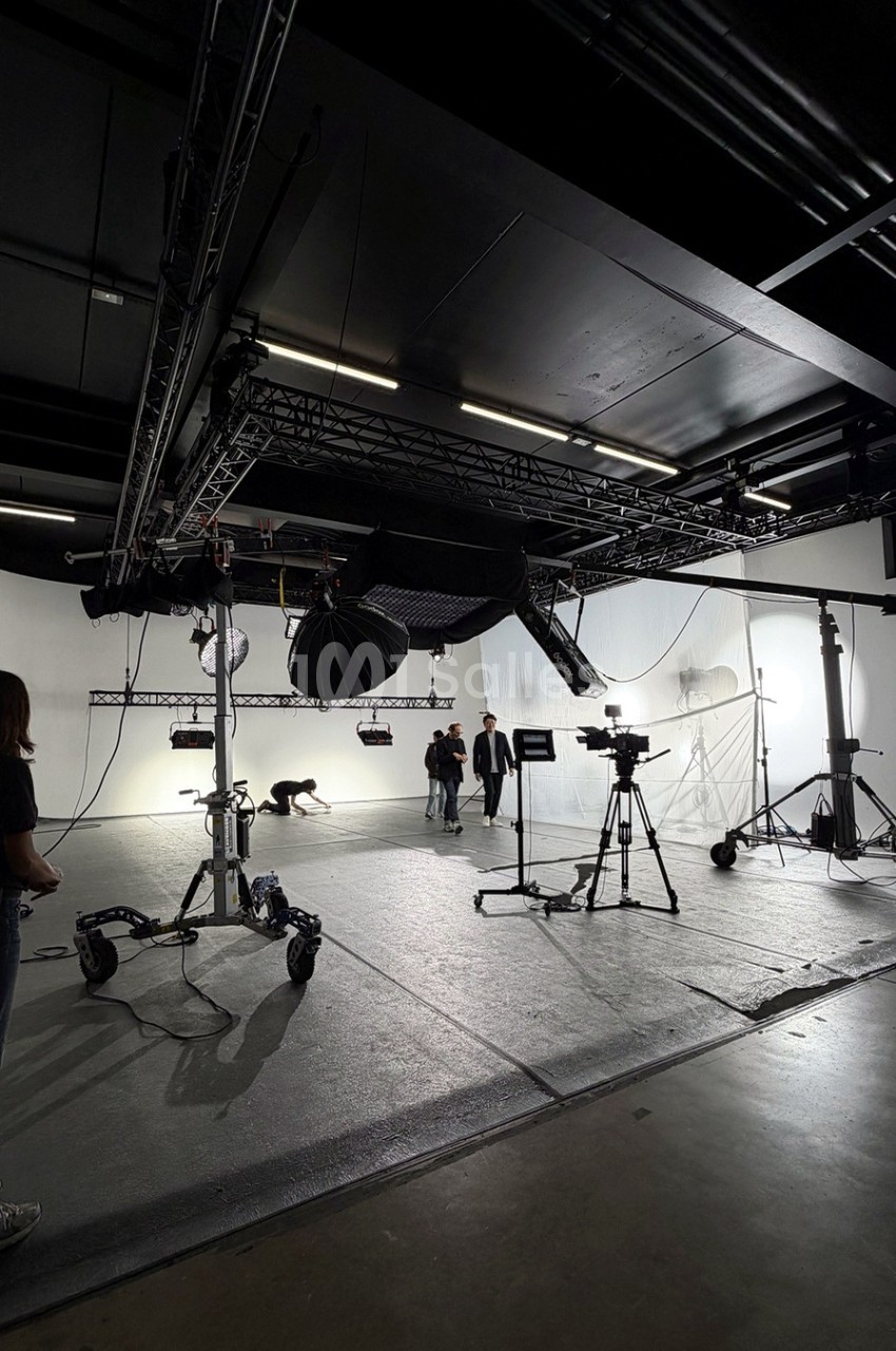 Plateau de tournage vide aux murs blancs et au sol gris, équipé de projecteurs suspendus à une structure en treillis, d'une caméra sur trépied et de pieds de lumière.