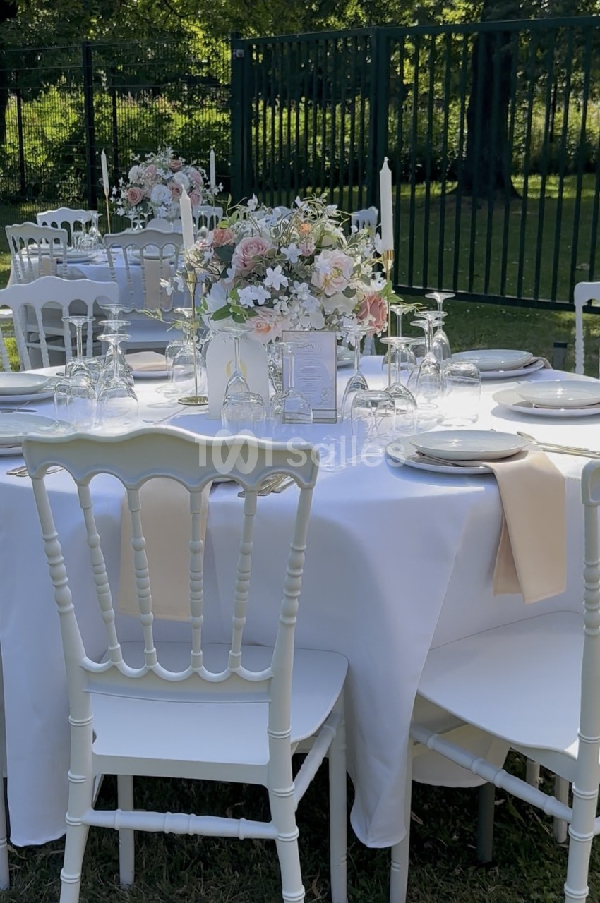 Tables rondes dressées en extérieur avec nappes blanches, chaises blanches et compositions florales roses et blanches.