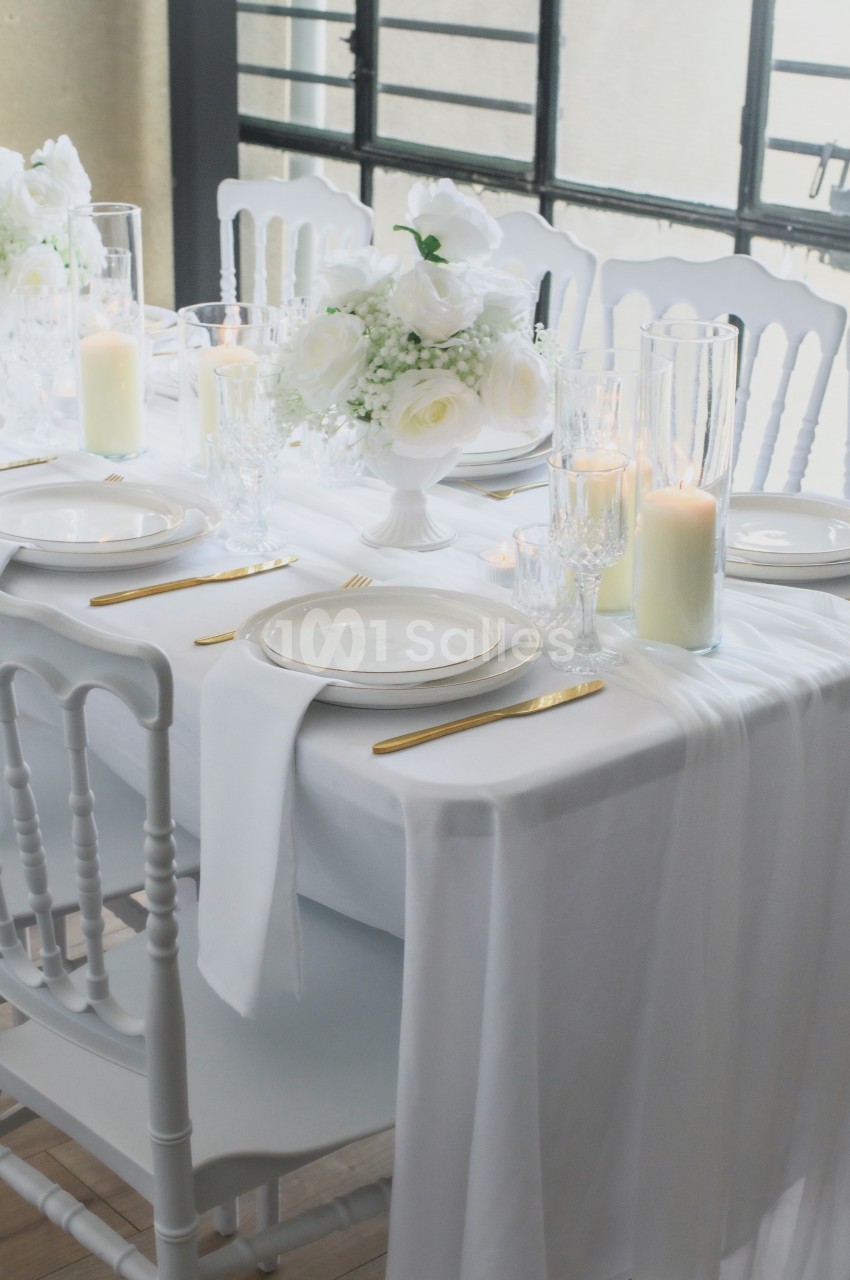Table ronde dressée en blanc et or avec chaises chiavari blanches, assiettes, verres et bougeoirs, fleurs blanches au centre.