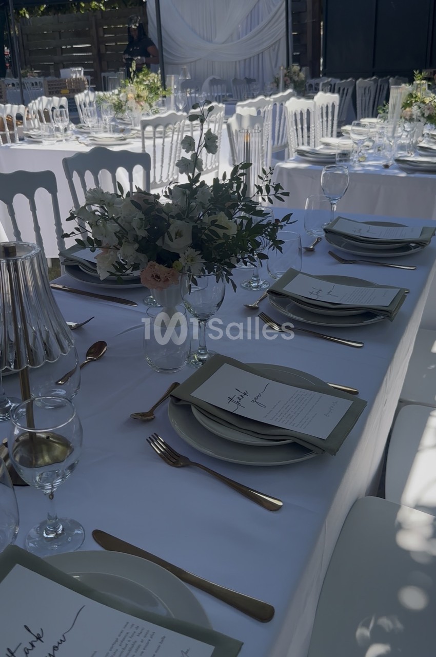 Salle dressée pour réception avec tables bleues, chaises blanches et centerpieces en fleurs.