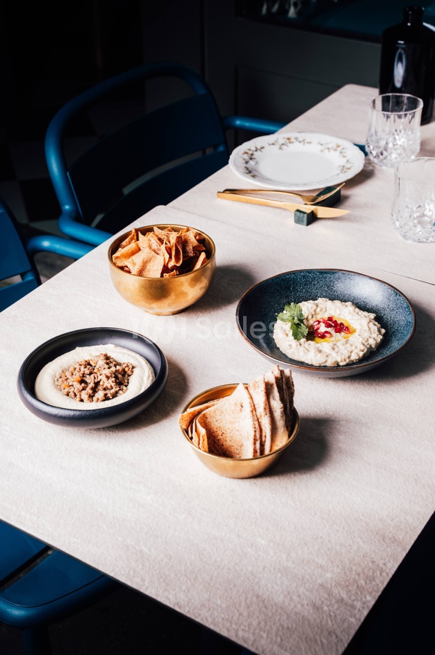 Table dressée avec mezzés - houmous, baba ganoush, chips et pain grillé dans bols dorés - chaise bleue en arrière-plan.