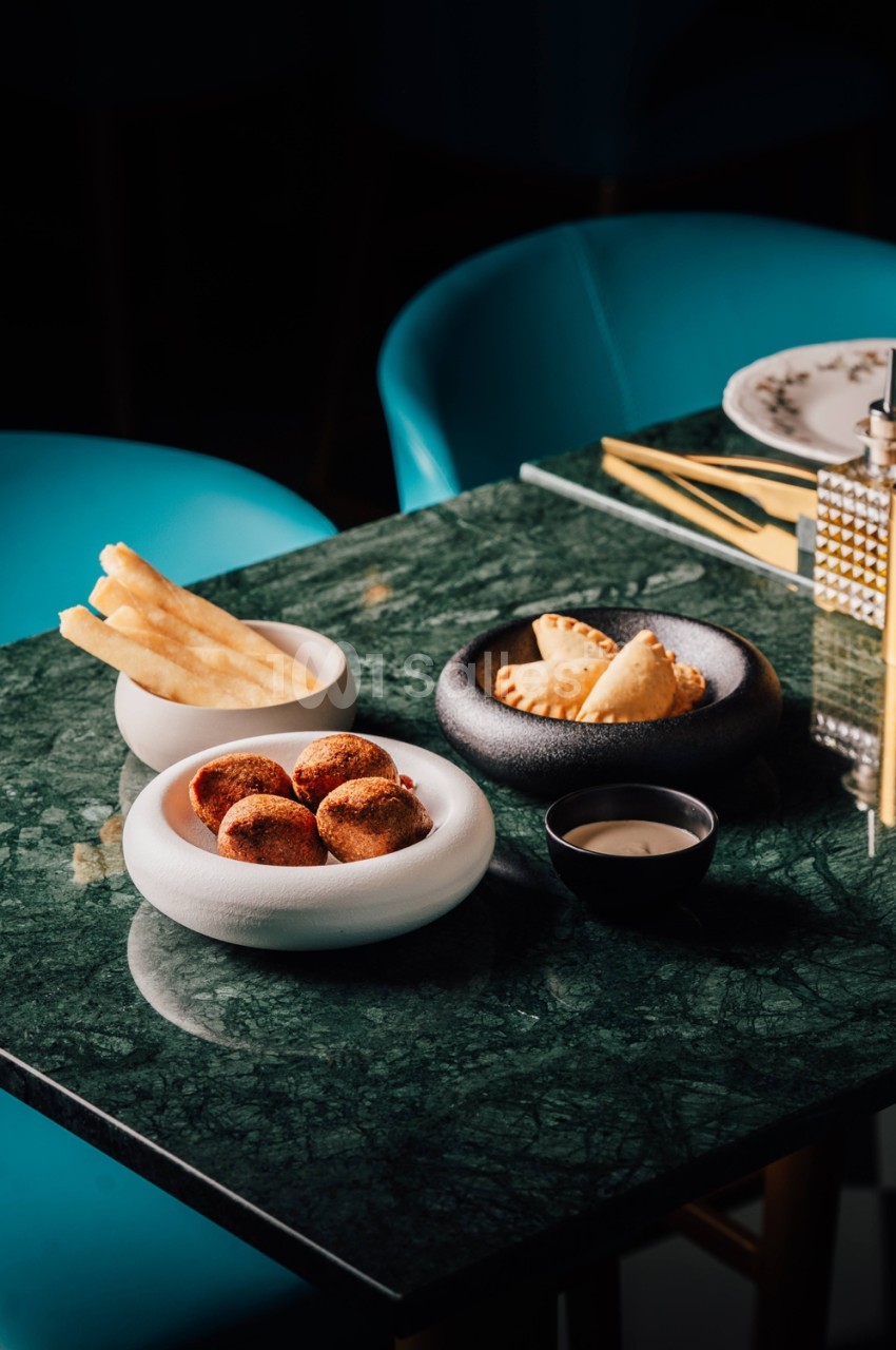 Table en marbre vert avec assiettes de frites, beignets et empanadillas, chaises turquoise en arrière-plan.