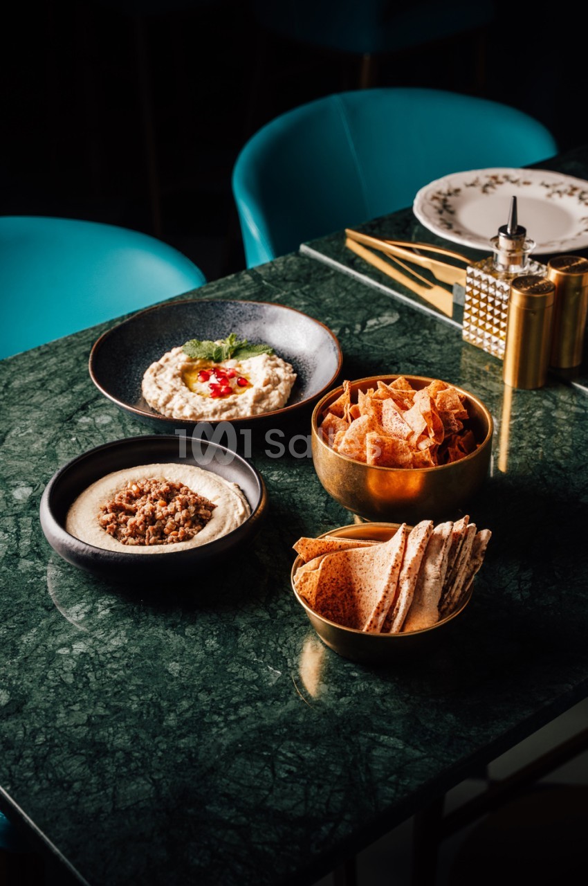 Table en marbre vert dressée avec assiettes de mezze - houmous, pains grillés, chips - et chaises turquoise en arrière-plan.