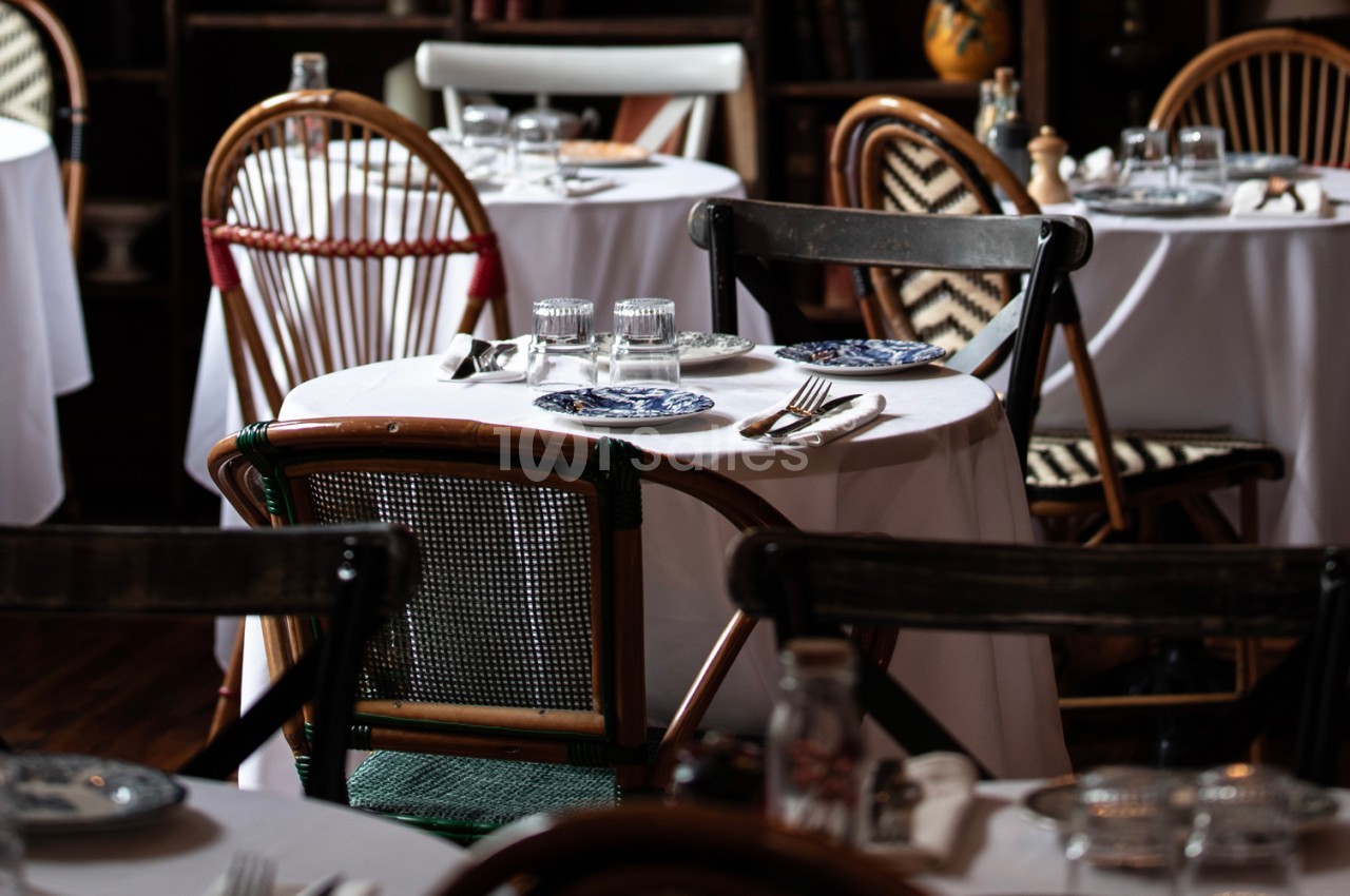 Salle de restaurant dressée avec tables rondes aux nappes blanches, chaises en bois et assiettes bleues.