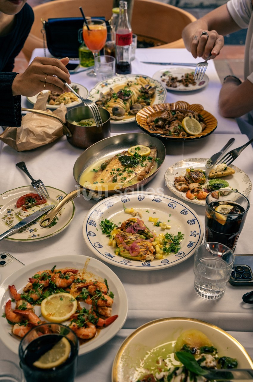 Table dressée avec convives mangeant des fruits de mer et poissons grillés, verres de boissons, paniers et assiettes variées.