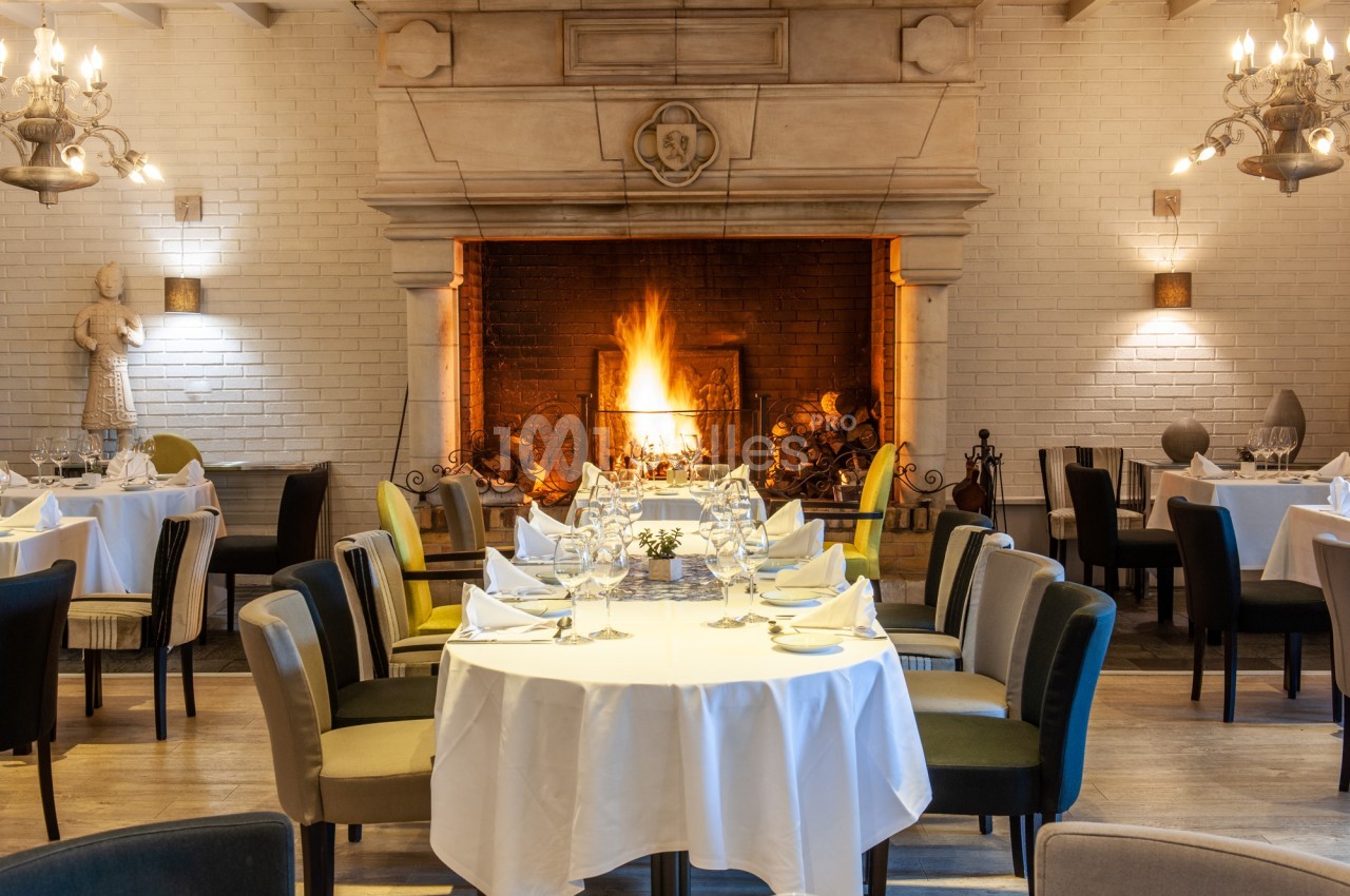 Salle de restaurant éclairée par des appliques, avec tables rondes dressées de nappes blanches, serviettes pliées et verres, devant une grande cheminée en pierre allumée.