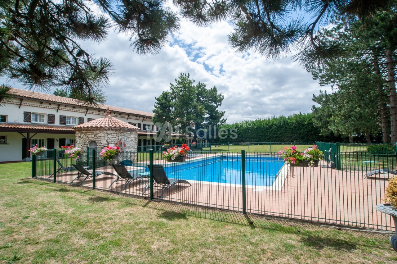 Piscine extérieure rectangulaire entourée d'une clôture verte, bordée de transats et de jardinières fleuries, près d'un bâtiment blanc à toiture de tuiles et de grands pins.