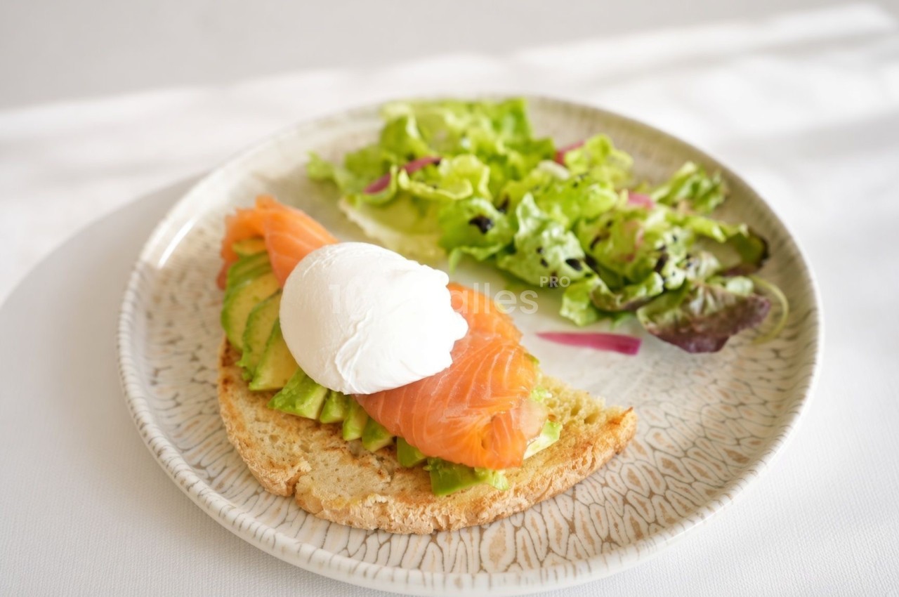 Tranche de pain grillé garnie d'avocat, de saumon fumé et d'un œuf poché, servie avec une salade verte sur une assiette en grès beige.
