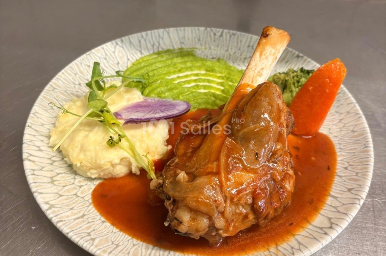 Jarret d'agneau braisé à la sauce tomatée, accompagné de purée, de courgettes vertes et d'une carotte, dressé sur une assiette texturée. Let me check: the image shows a braised lamb shank with a red sauce, mashed potatoes, green zucchini slices (looks