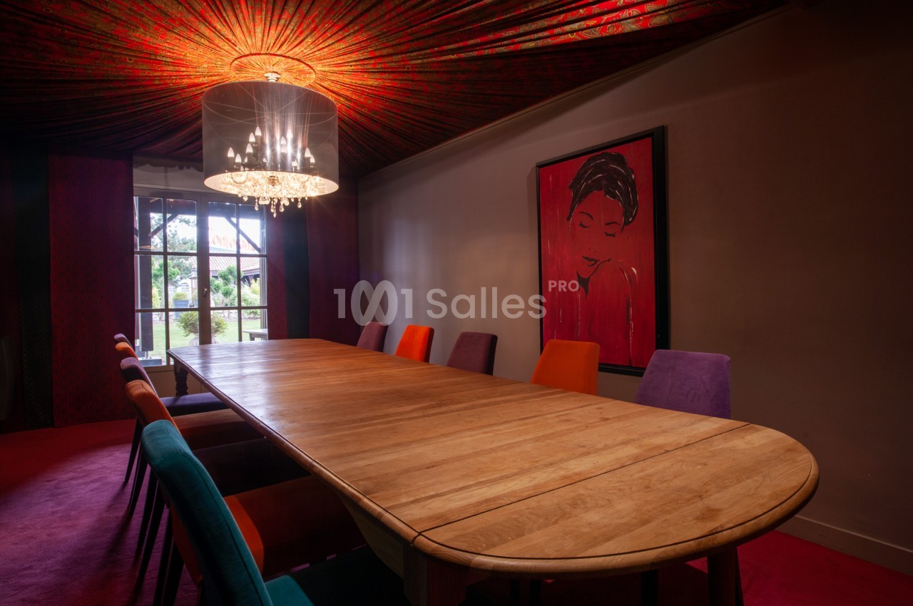 Salle de réunion aux murs et chaperon rouges, avec une grande table en bois massif entourée de chaises colorées et un lustre suspendu. Let me check constraints: one sentence, under 125 chars? Count: "Salle de réunion aux murs et chaperon rouges" - wrong