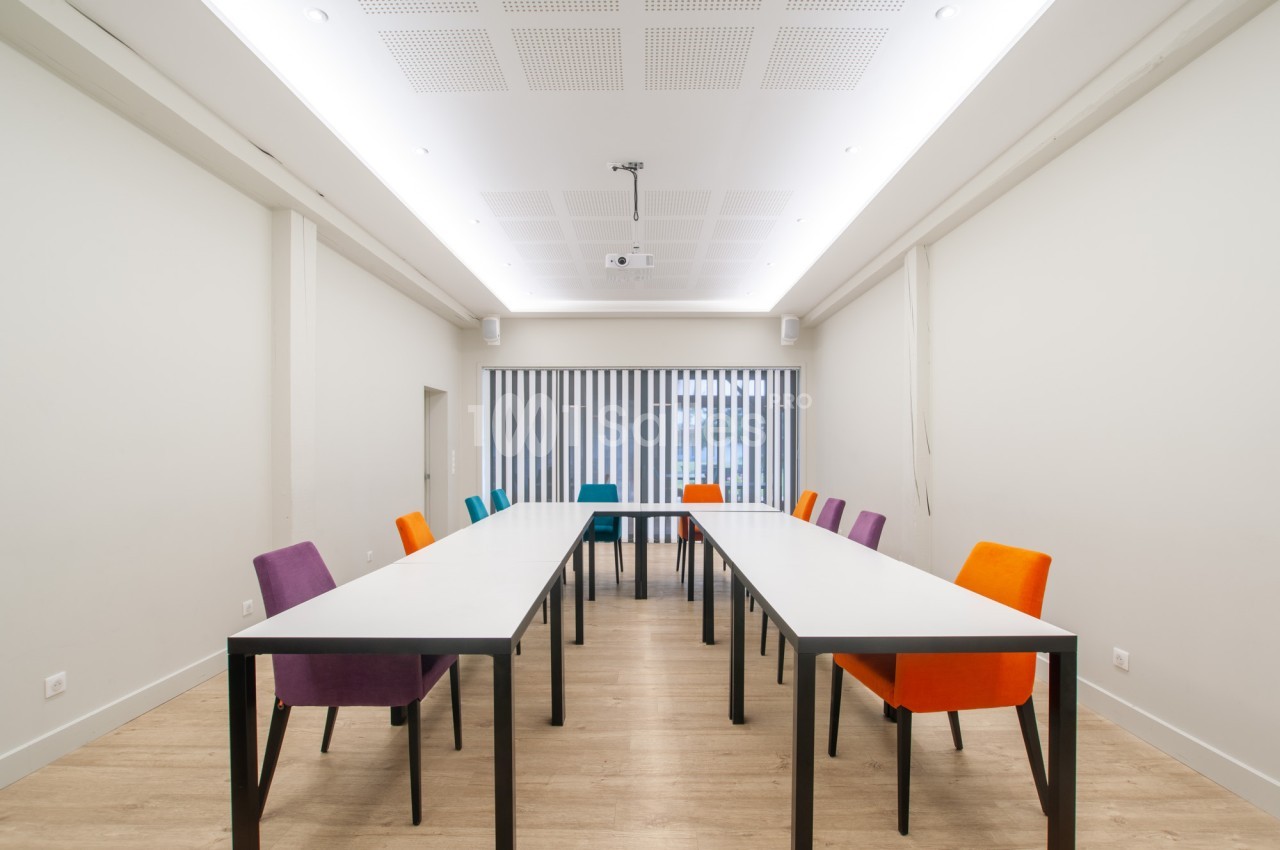 Salle de réunion aux murs blancs, avec deux longues tables blanches en U et des chaises aux couleurs variées (violet, orange, bleu), vidéoprojecteur au plafond. Let me check length: roughly 165 characters, too long. Need ≤125. Salle sans fenêtre? There