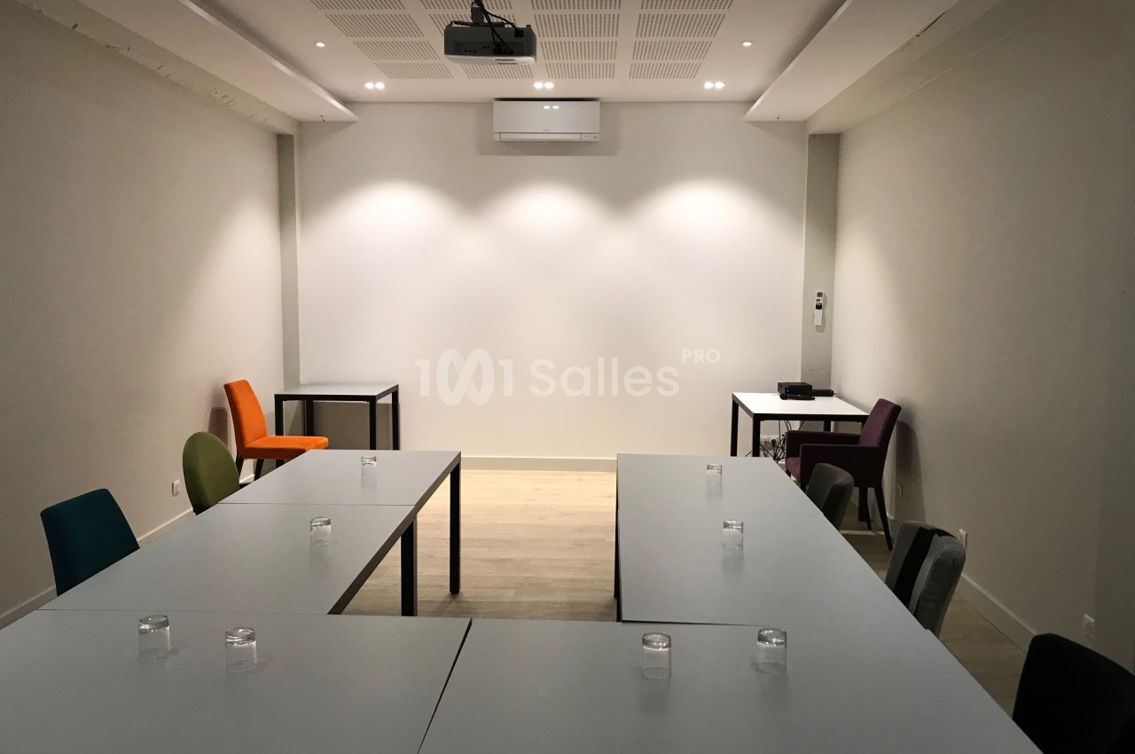 Salle de réunion aménagée en U avec tables grises, chaises colorées, vidéoprojecteur au plafond et mur blanc éclairé en fond.