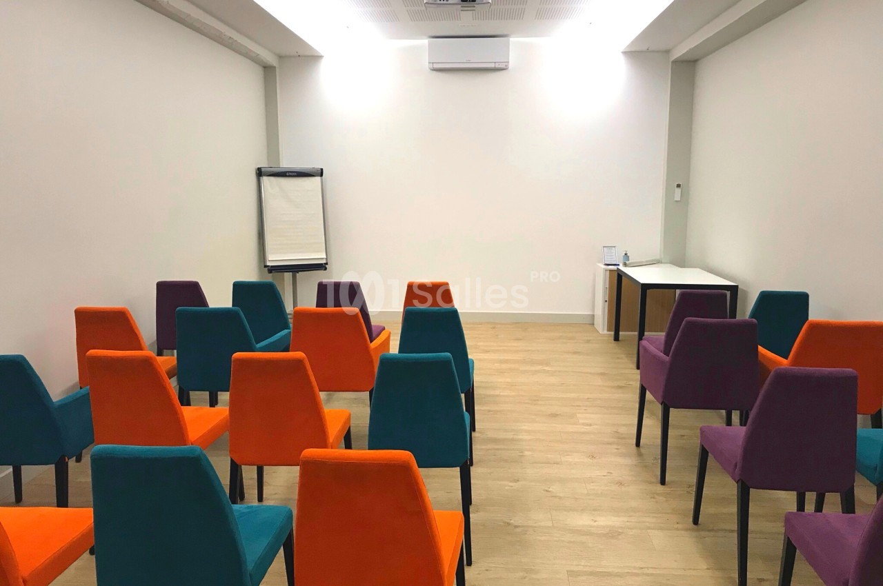 Salle de réunion aux murs blancs, avec des rangées de chaises aux couleurs orange, bleu canard et violet, disposées face à un tableau de conférence sur chevalet. Let me check length: about 150 characters - too long. Need ≤125. Salle de réunion vide aux
