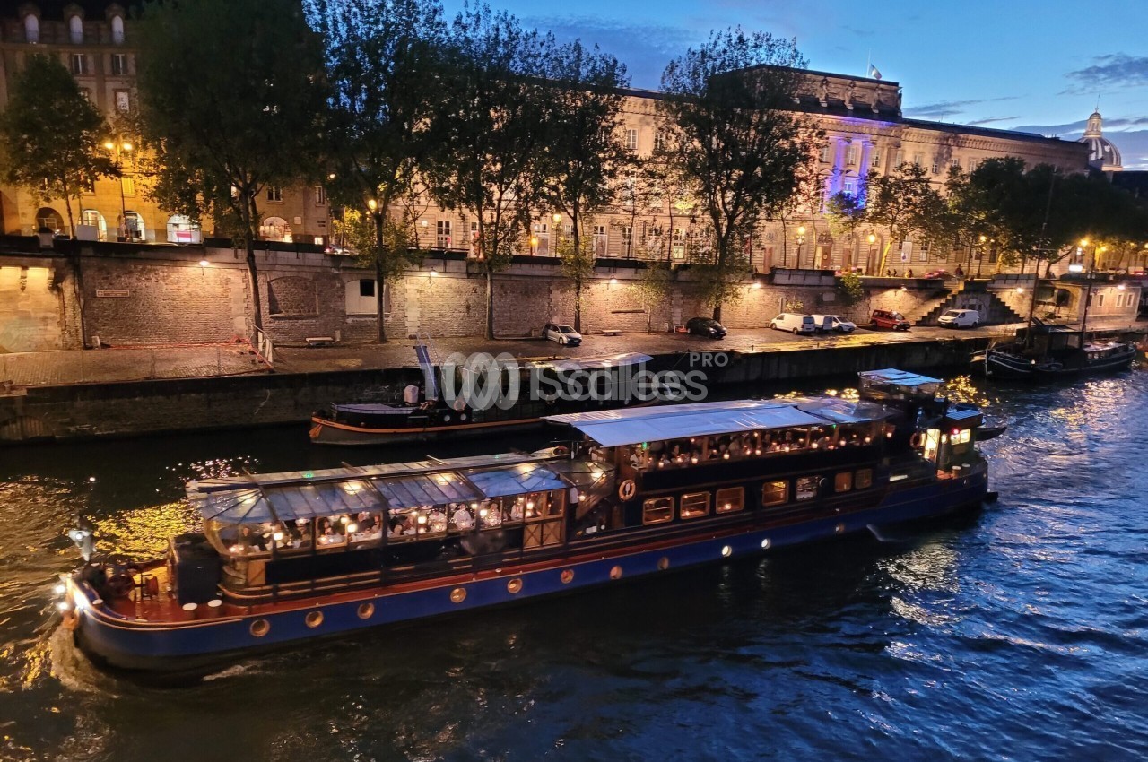 Péniche restaurant éclairée de nuit sur la Seine, avec des convives attablés sous une verrière, à côté de quais en pierre et d'arbres.