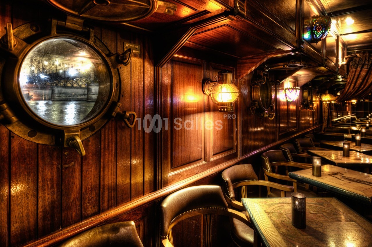Intérieur de bateau-restaurant aux boiseries sombres, avec un hublot à gauche, des appliques en laiton et des tables alignées le long d'une banquette de fauteuils en cuir.