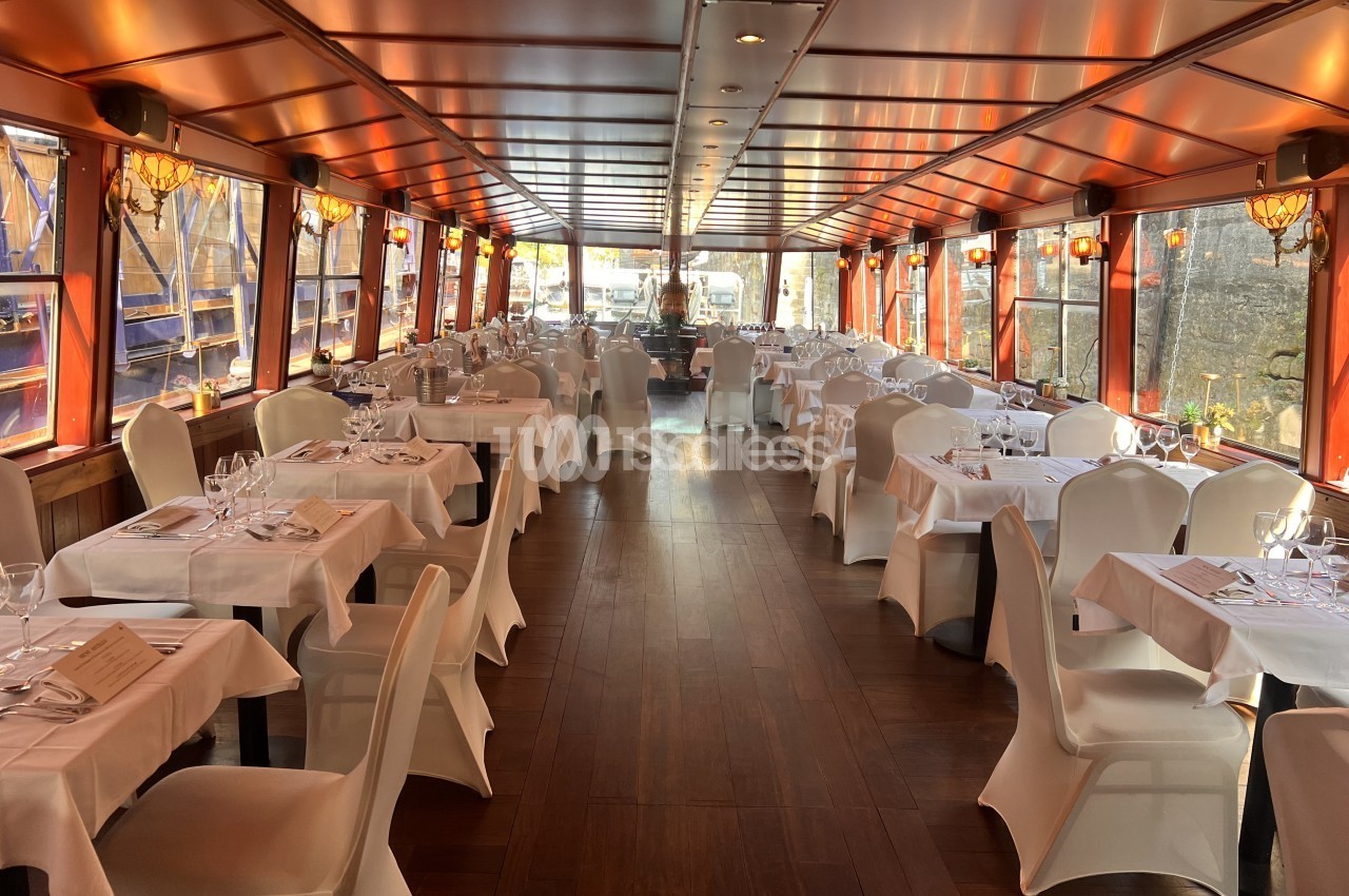 Salle à bord d'un bateau aux tables dressées de nappes et housses de chaises blanches, le long de grandes baies vitrées.